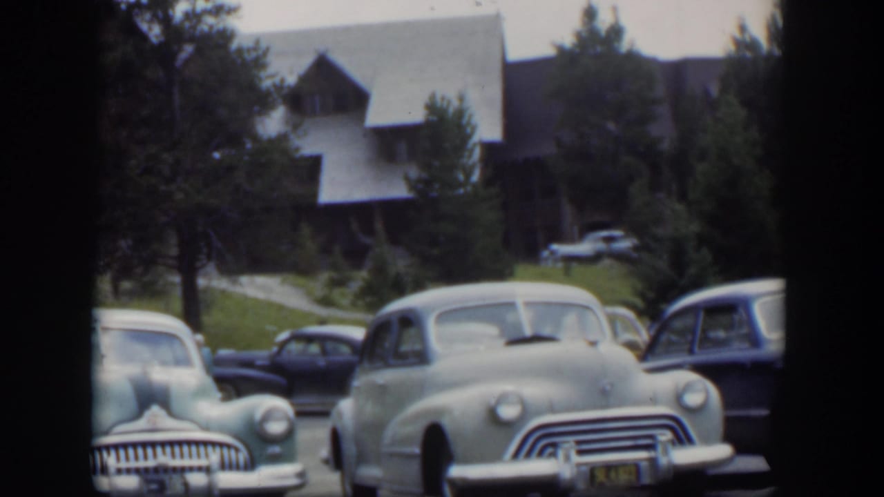 1951 Wyoming home-movie frame with a pale rounded car centered among parked vehicles near a steep-roof lodge and trees.