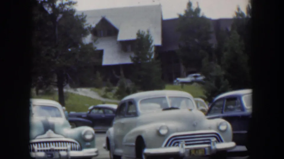 1951 Wyoming home-movie frame with a pale rounded car centered among parked vehicles near a steep-roof lodge and trees.