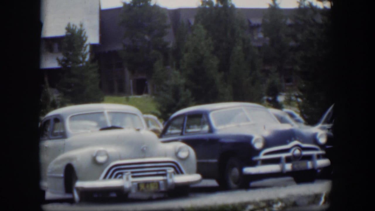 1951 Wyoming frame showing a pale rounded car beside a darker sedan, with lodge structures and evergreens in the background.
