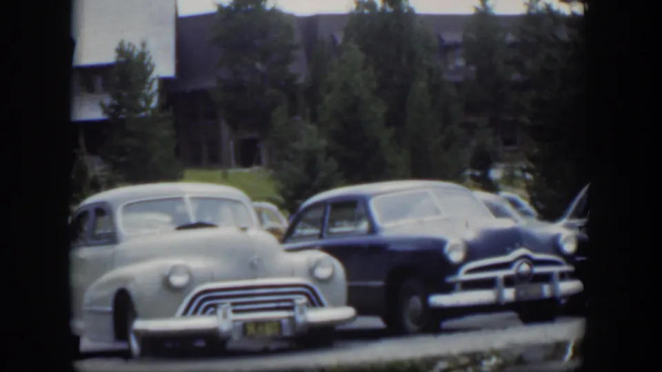 1951 Wyoming frame showing a pale rounded car beside a darker sedan, with lodge structures and evergreens in the background.