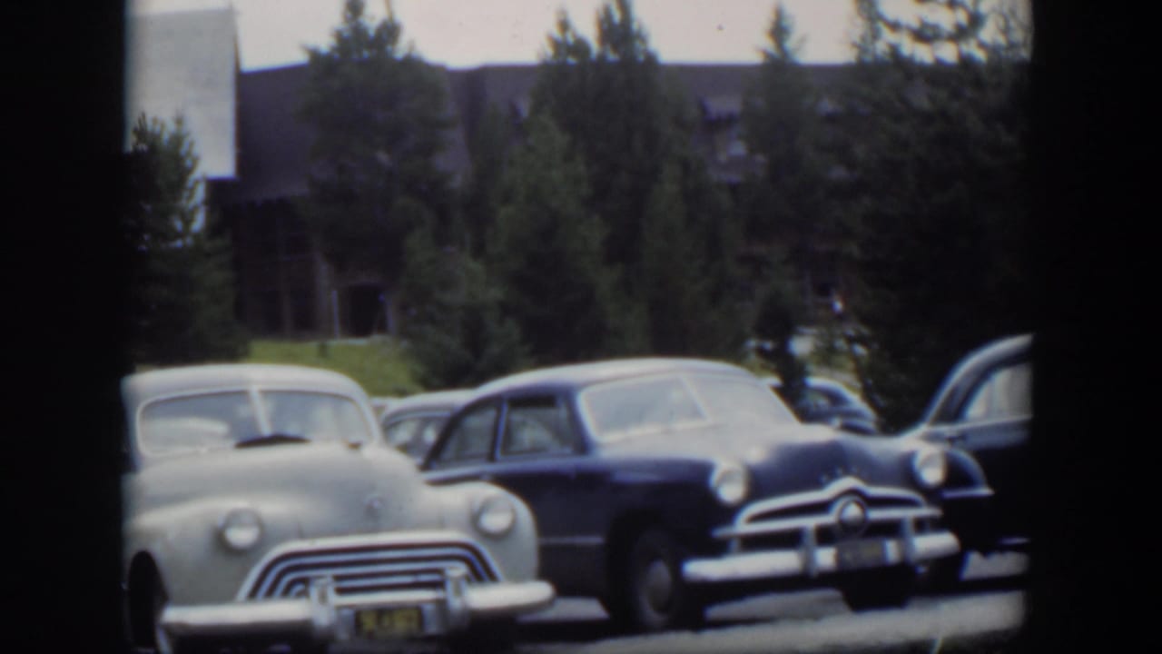 Classic cars parked in front of a lodge with evergreen trees behind them.