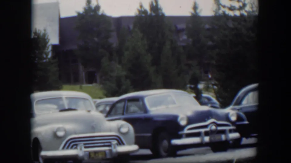1951 Wyoming 8mm frame showing light and dark mid-century cars parked before a lodge-like building and tall evergreens.