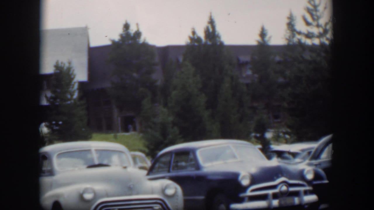 1951 Wyoming home-movie frame with a pale sedan and dark car in a lodge parking area under soft overcast light.