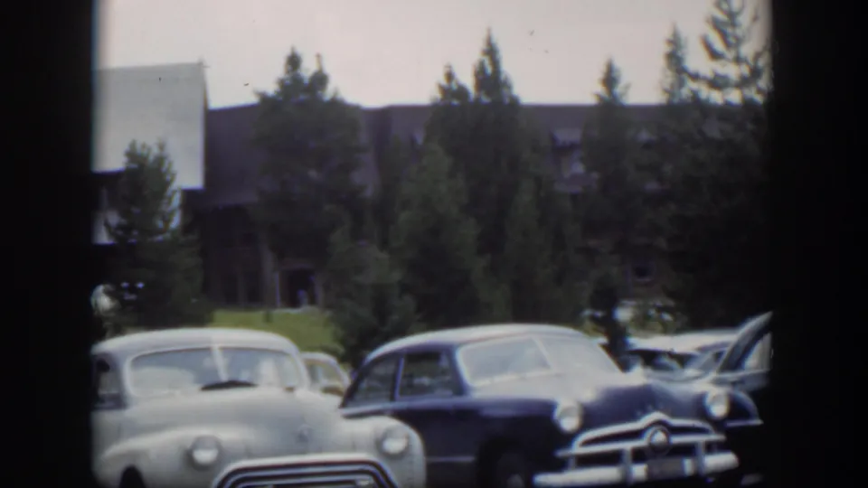 1951 Wyoming home-movie frame with a pale sedan and dark car in a lodge parking area under soft overcast light.