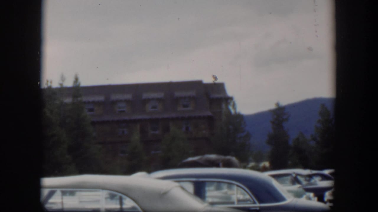 1951 Wyoming frame showing a lodge-like building beyond parked cars, with evergreens and a dark mountain slope in the distance.