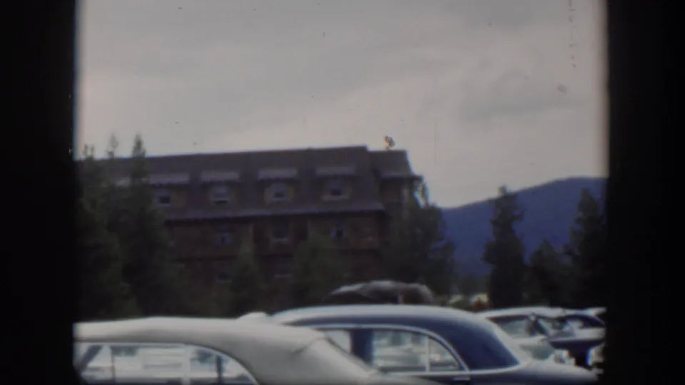 1951 Wyoming frame showing a lodge-like building beyond parked cars, with evergreens and a dark mountain slope in the distance.