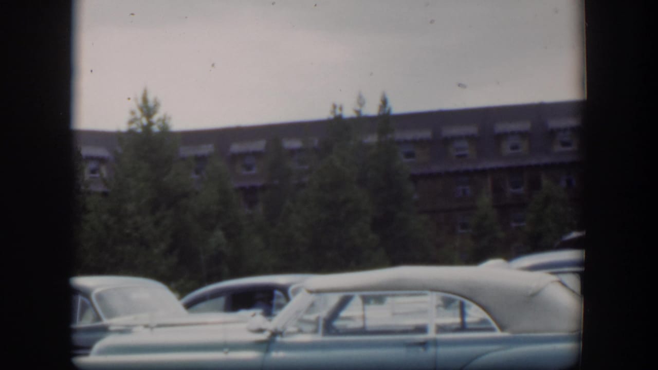 1951 Wyoming home-movie frame with parked mid-century cars partly obscuring pine trees and a large brown building with dormer windows.