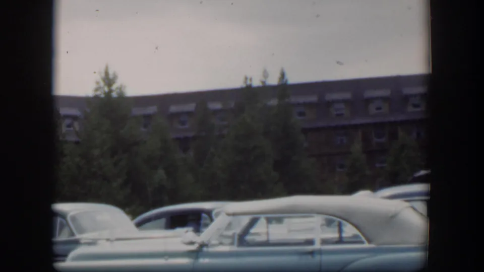 1951 Wyoming home-movie frame with parked mid-century cars partly obscuring pine trees and a large brown building with dormer windows.