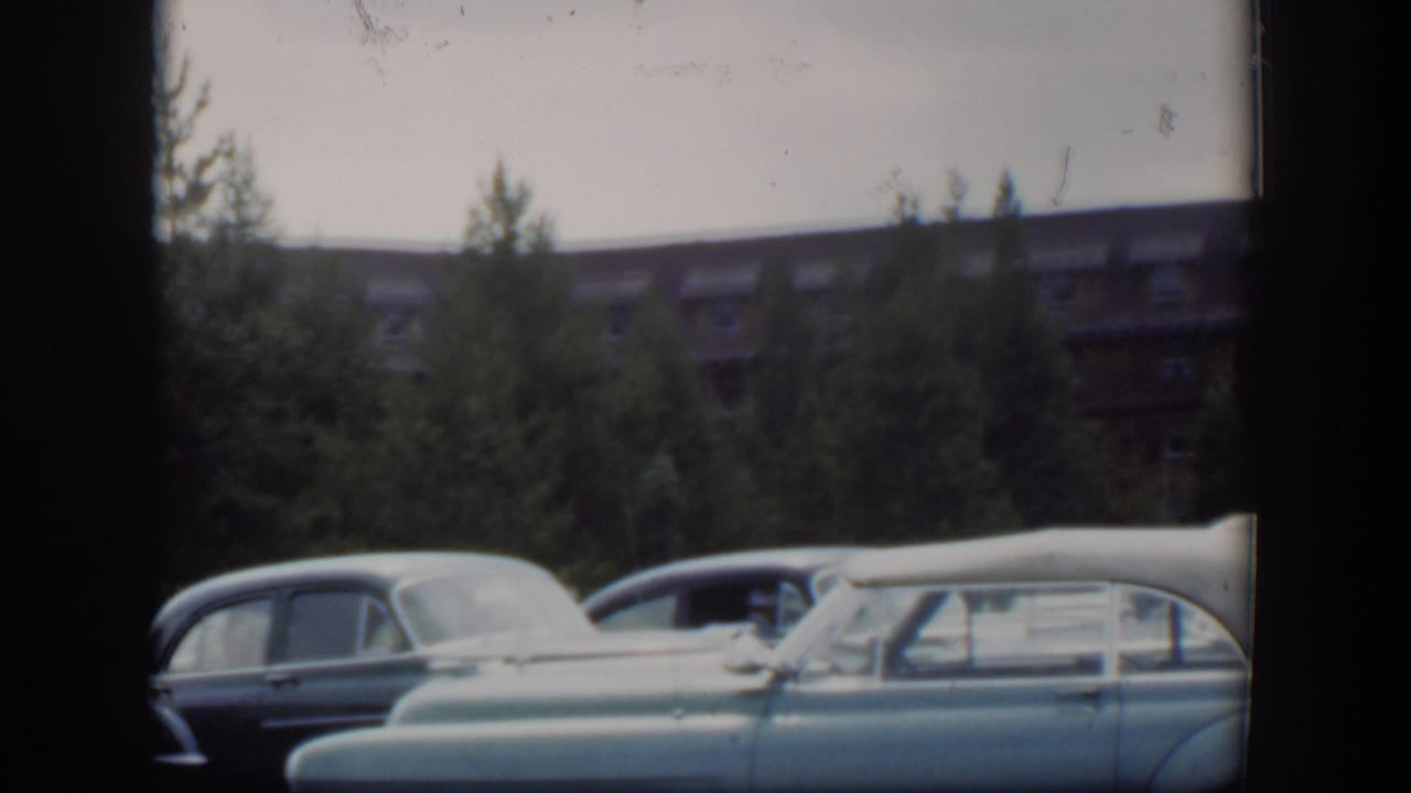 1951 Wyoming 8mm frame showing several parked classic cars in the foreground with trees and a dormer-windowed brown building behind them.