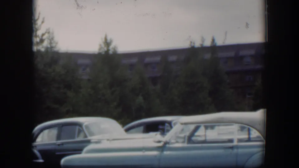 1951 Wyoming 8mm frame showing several parked classic cars in the foreground with trees and a dormer-windowed brown building behind them.