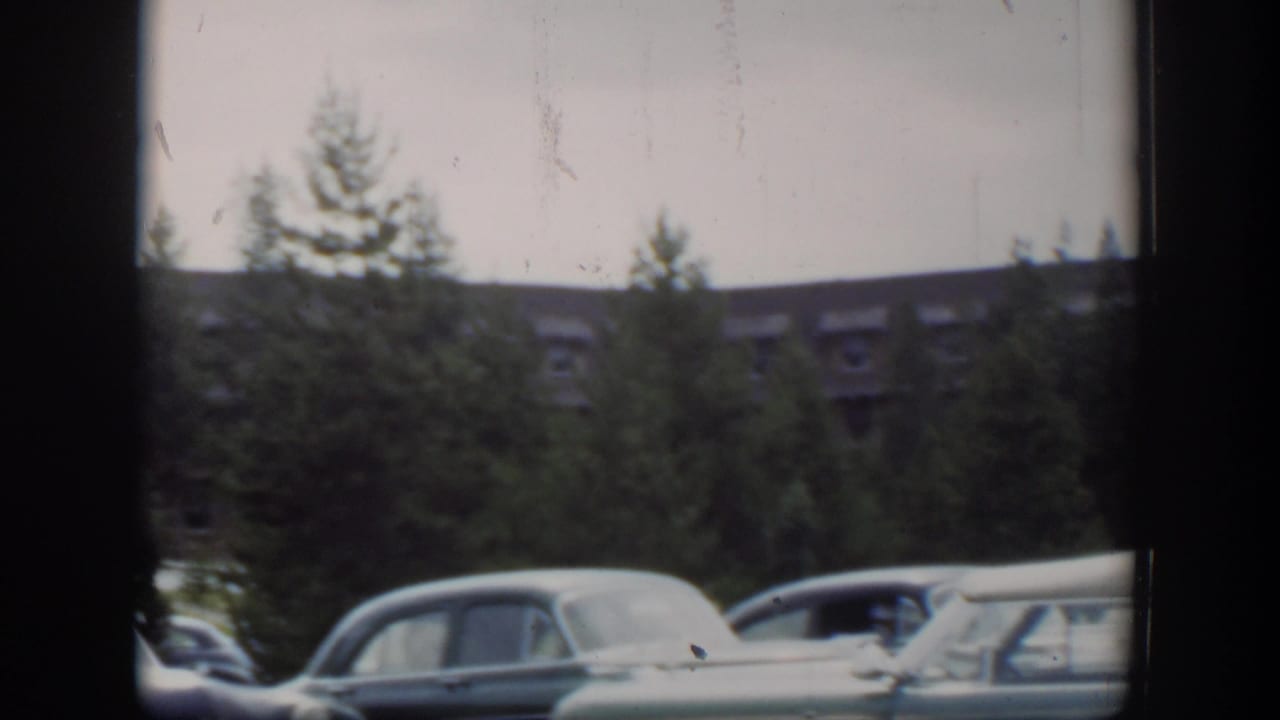 Late-frame 1951 Wyoming view of parked mid-century cars beneath pine trees, with the brown dormer-roofed building receding in the background.