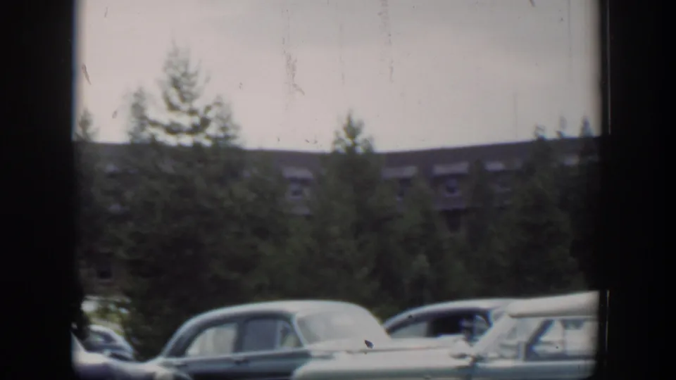 Late-frame 1951 Wyoming view of parked mid-century cars beneath pine trees, with the brown dormer-roofed building receding in the background.