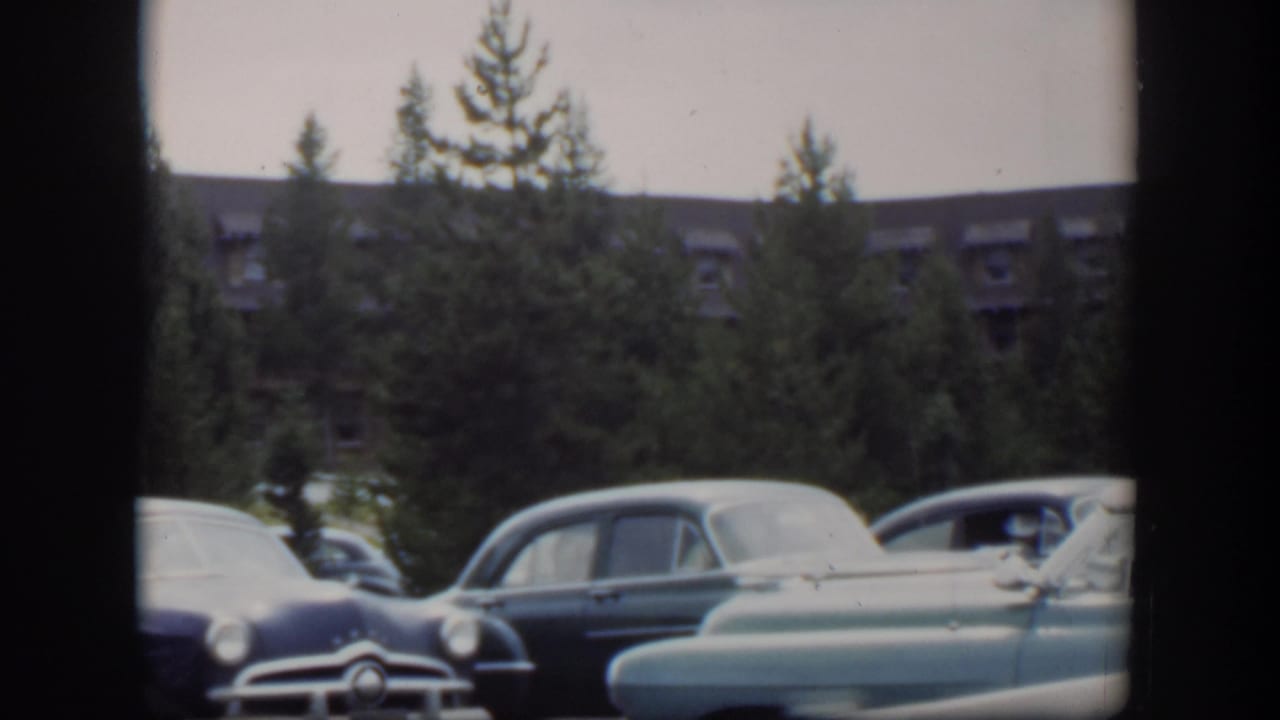 1951 Wyoming 8mm frame showing rounded mid-century cars parked near trees, with a long distant building partly visible beyond them.