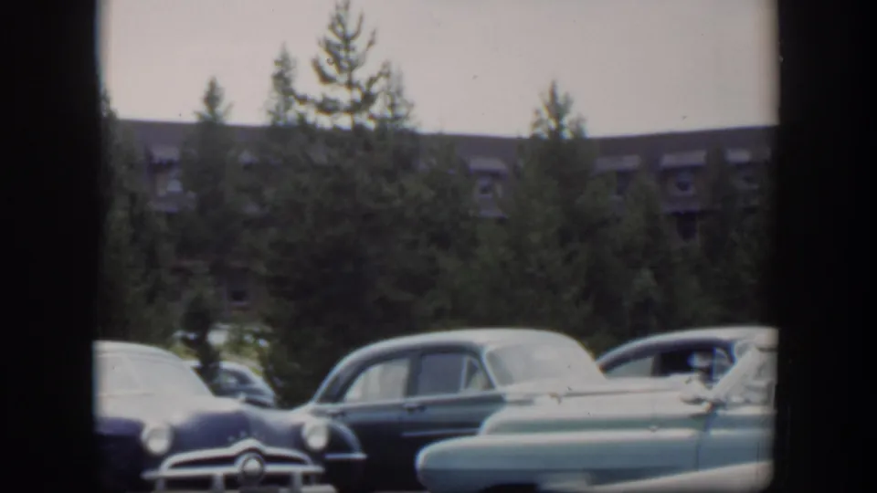 1951 Wyoming 8mm frame showing rounded mid-century cars parked near trees, with a long distant building partly visible beyond them.