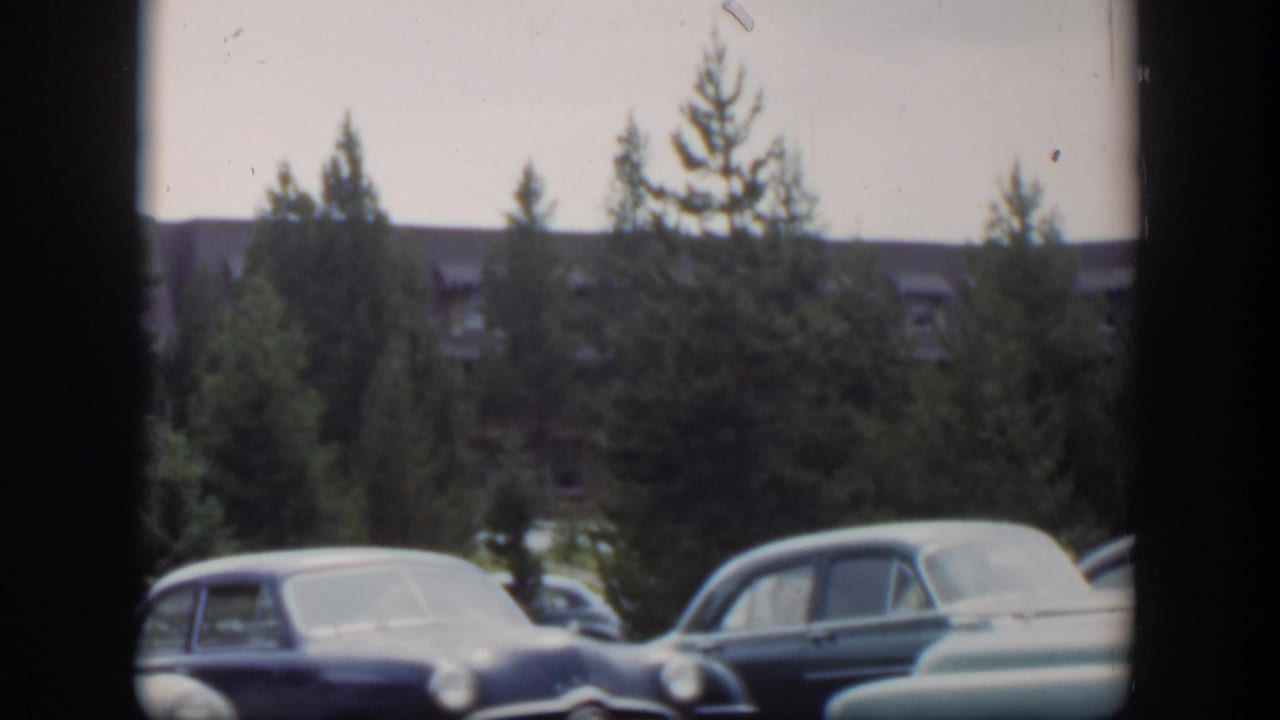 Parked sedans and a light car in the foreground with trees and a building behind.