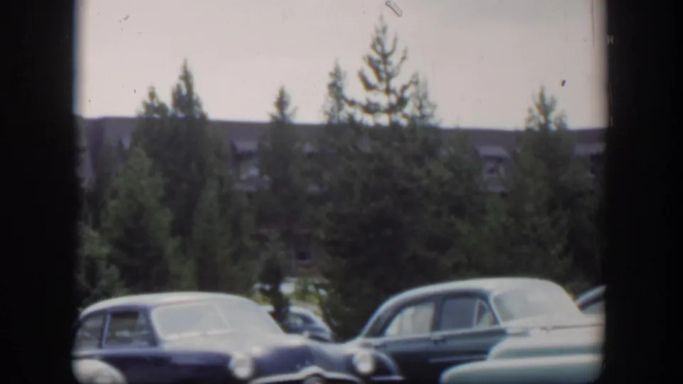 1951 Wyoming frame with several early-1950s automobiles in a parking area, backed by evergreens and a long building.