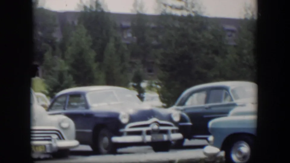 1951 Wyoming frame with a light car moving across the foreground while classic sedans remain parked near trees and a distant building.