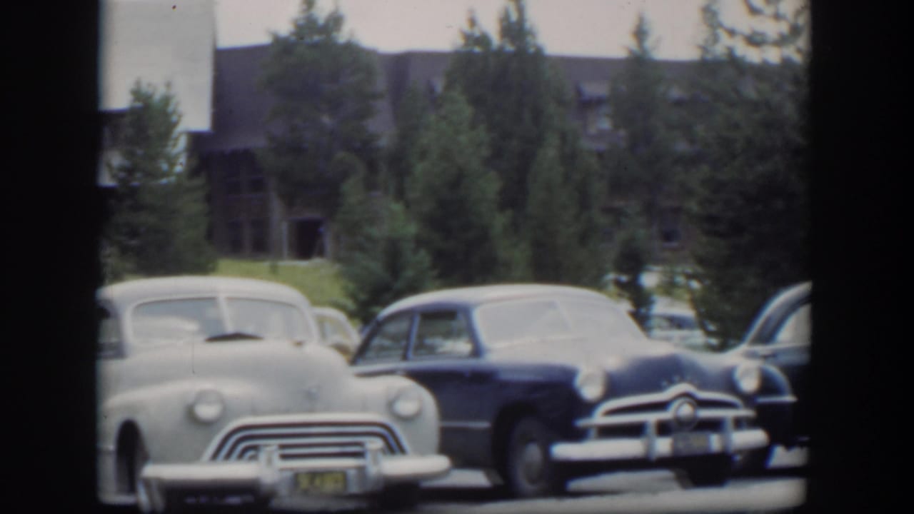 Light gray sedan and dark blue car parked before a lodge with evergreen trees.