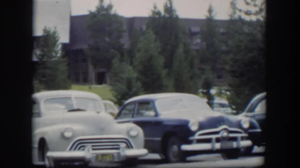 1951 Wyoming 8mm frame showing two early 1950s cars in a parking lot near a rustic lodge and evergreens.