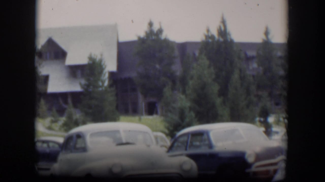 Early 1950s cars appear in a Wyoming lodge parking area, with evergreens and dark frame edges from the 8mm scan.