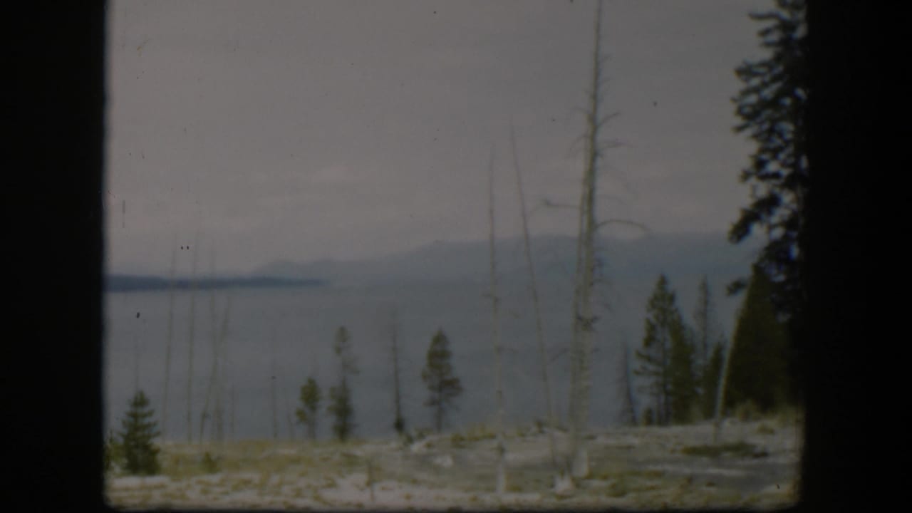 1951 Wyoming 8mm frame showing calm water, distant hills, pale rocky ground, and scattered trees in the foreground.