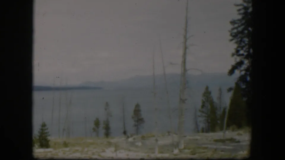 1951 Wyoming 8mm frame showing calm water, distant hills, pale rocky ground, and scattered trees in the foreground.
