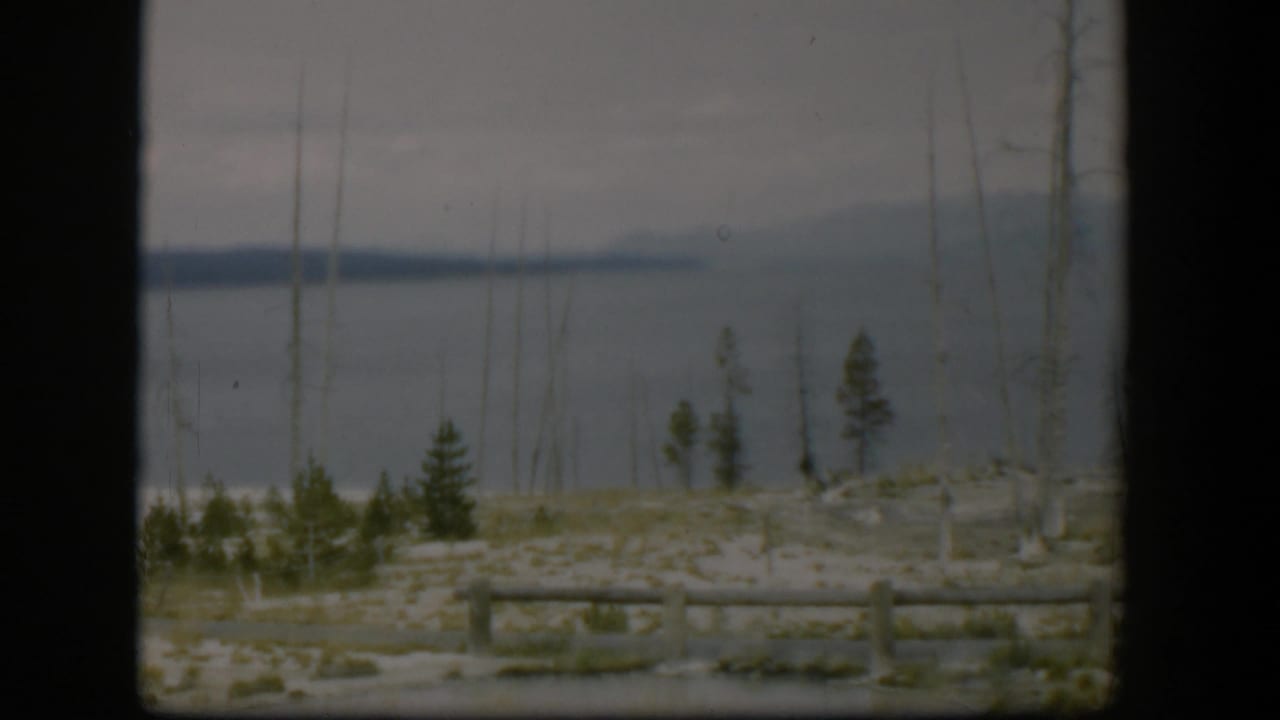 1951 Wyoming 8mm frame with calm water, a dark distant shore, sparse foreground trees, pale ground, and log fencing.