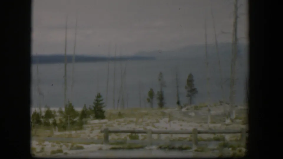 1951 Wyoming 8mm frame with calm water, a dark distant shore, sparse foreground trees, pale ground, and log fencing.