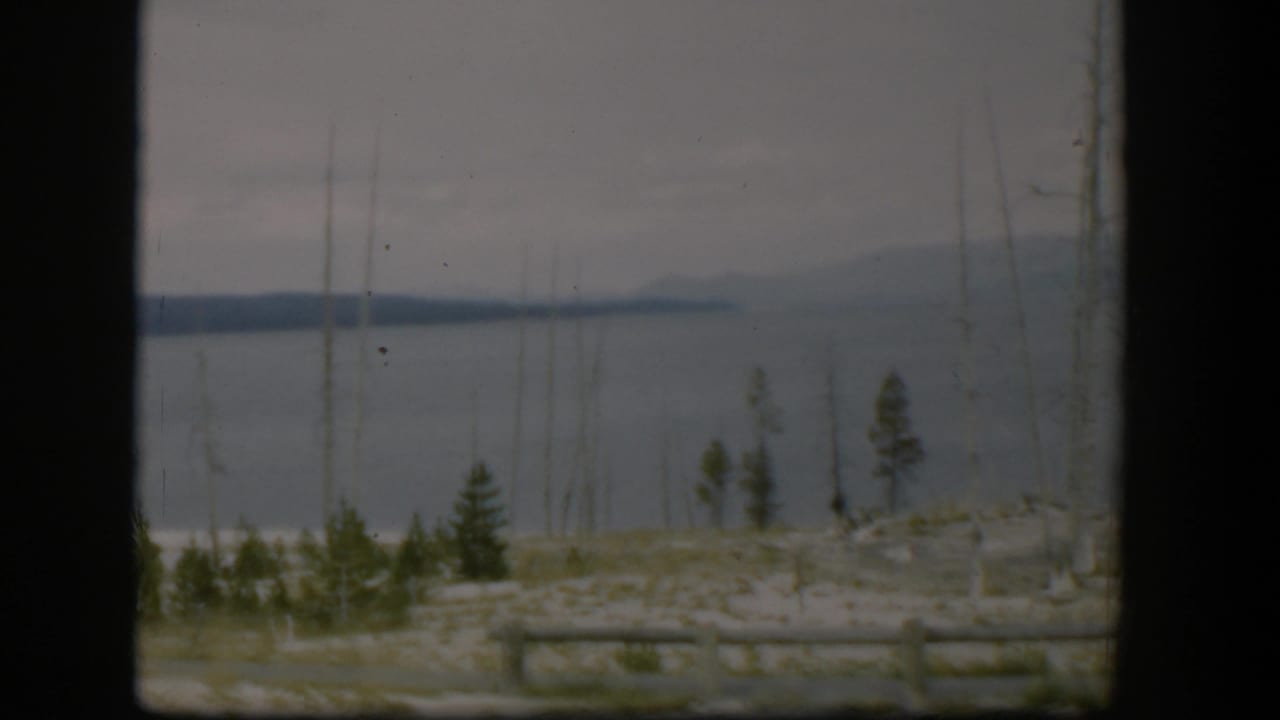 1951 Wyoming home-movie frame showing a gray water view with distant land, sparse trees, and roadside log fencing.