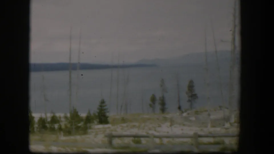 1951 Wyoming home-movie frame showing a gray water view with distant land, sparse trees, and roadside log fencing.