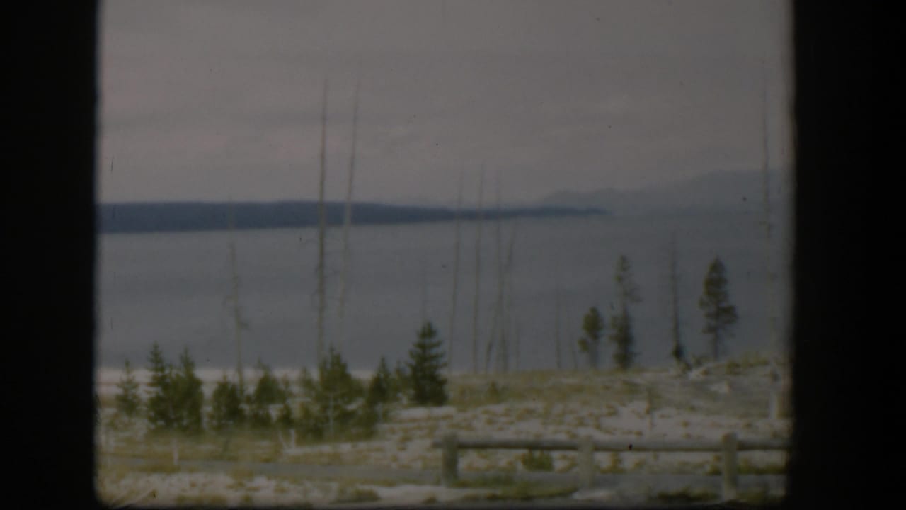Large lake seen through a dark window frame with evergreens, bare trunks, rail fence, and distant hills.