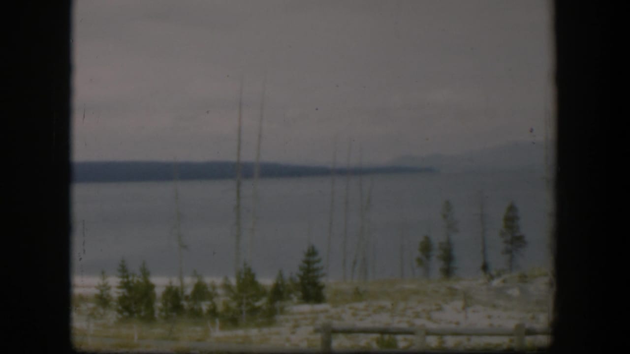 1951 Wyoming lake view framed by dark vertical edges, with low hills across the water, small evergreens, dead trunks, and rail fencing.