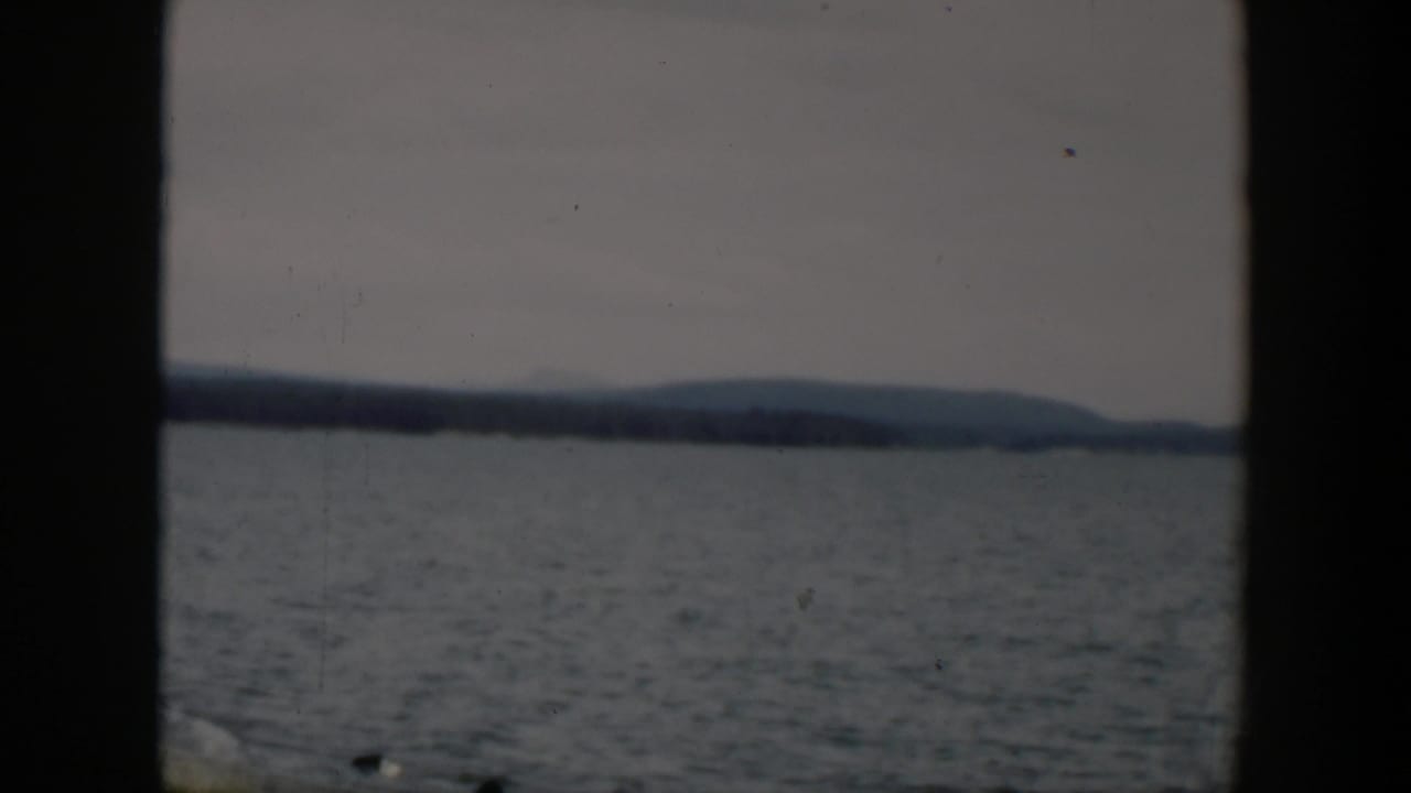 Lake water with distant tree-lined shore and low hills, seen through dark 8mm frame borders.