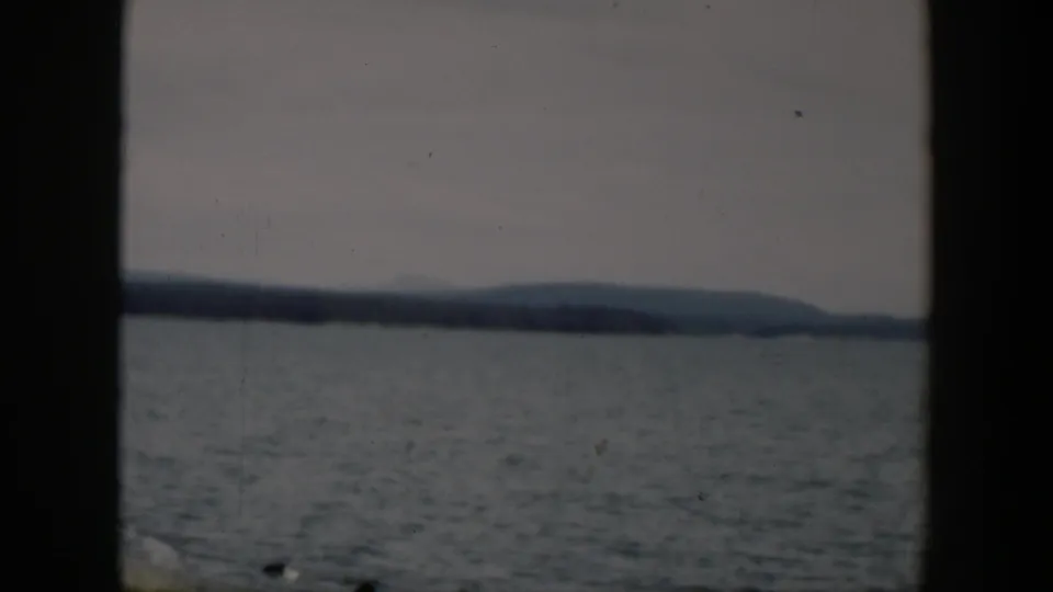 1951 Wyoming 8mm frame showing rippled lake water, a distant wooded shoreline, low hills, and a gray-blue sky.