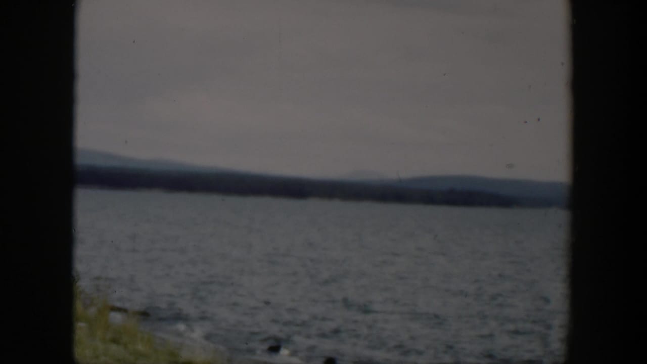 1951 Wyoming 8mm view of rippled lake water, far trees, low hills, muted sky, and a small strip of grassy shore.