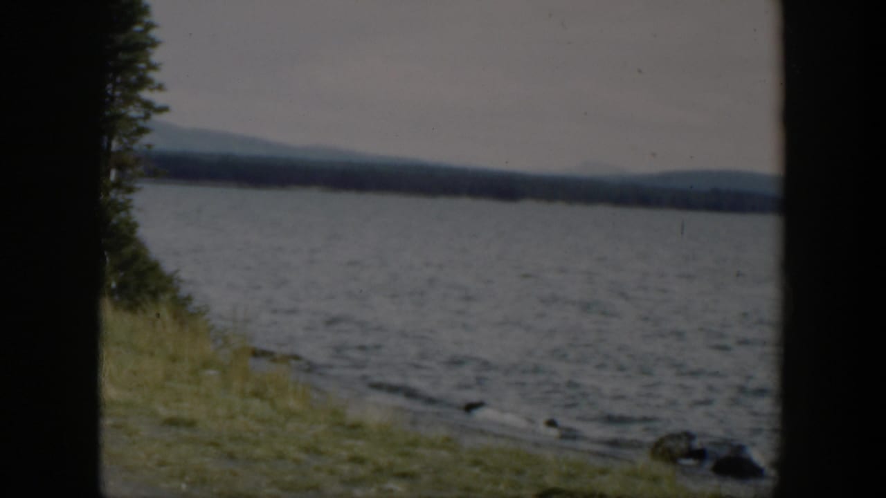 1951 Wyoming 8mm frame showing rippled lake water, small shoreline rocks, grass, and distant hills in soft focus.