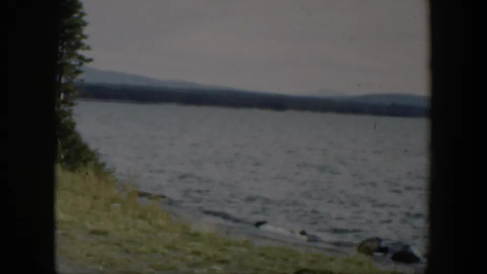 1951 Wyoming 8mm frame showing rippled lake water, small shoreline rocks, grass, and distant hills in soft focus.