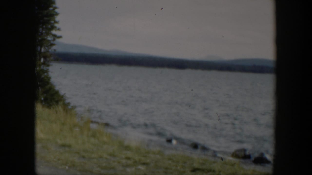 1951 Wyoming 8mm frame showing a grassy lakeshore, rippled water, evergreen edge, rocks, and distant mountain ridges.
