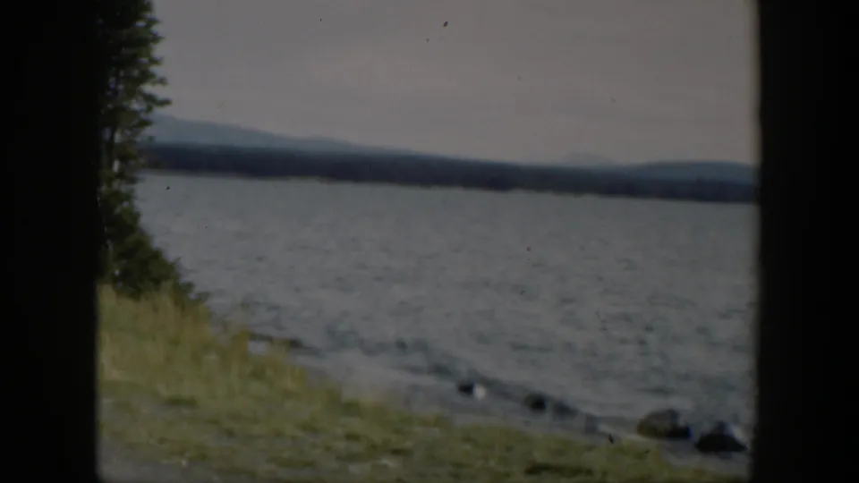1951 Wyoming 8mm frame showing a grassy lakeshore, rippled water, evergreen edge, rocks, and distant mountain ridges.