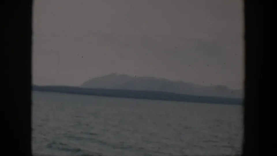 1951 Wyoming 8mm frame showing gray water, a far shoreline, and a soft mountain silhouette beneath a cloudy sky.