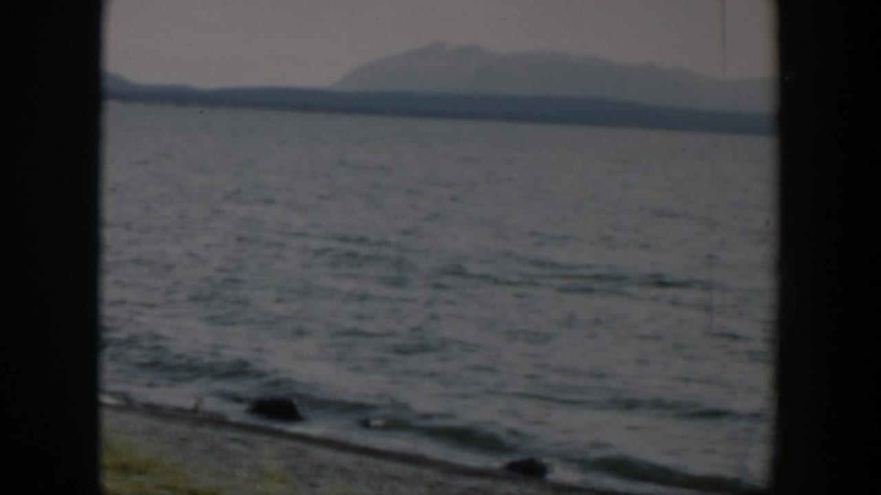1951 Wyoming 8mm frame showing gentle lake waves, a stony bank, low grass, and a hazy mountain range across the water.