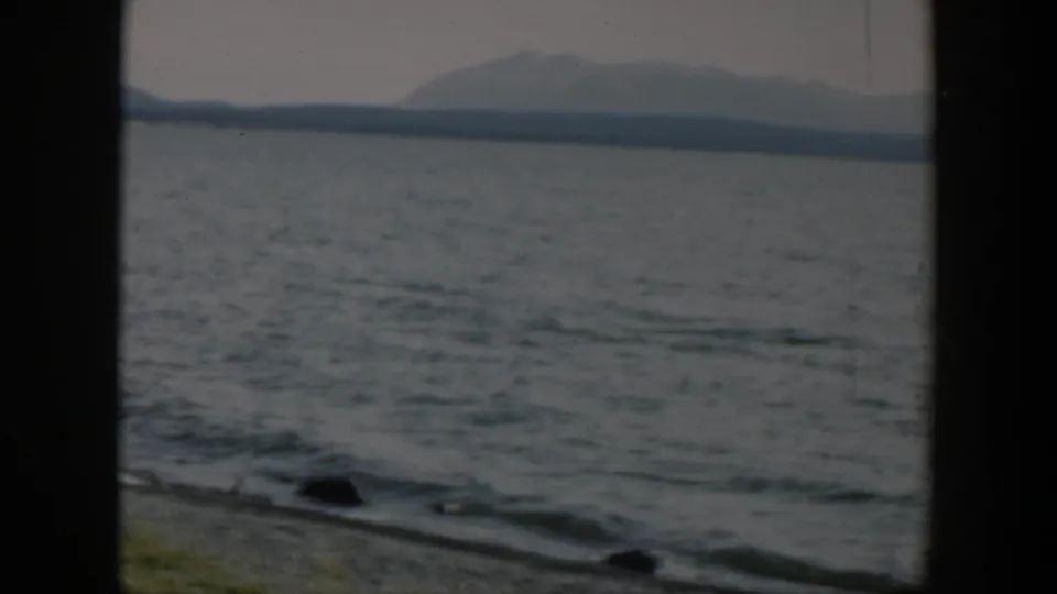 1951 Wyoming 8mm frame showing gentle lake waves, a stony bank, low grass, and a hazy mountain range across the water.