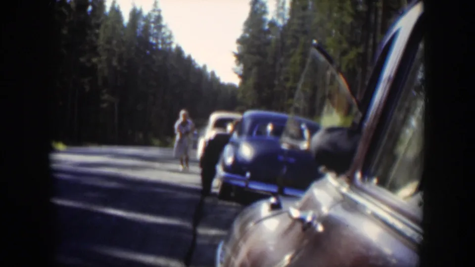 Mid-century Wyoming home movie frame with cars pulled over, a woman on the road, and a bear near the vehicles.