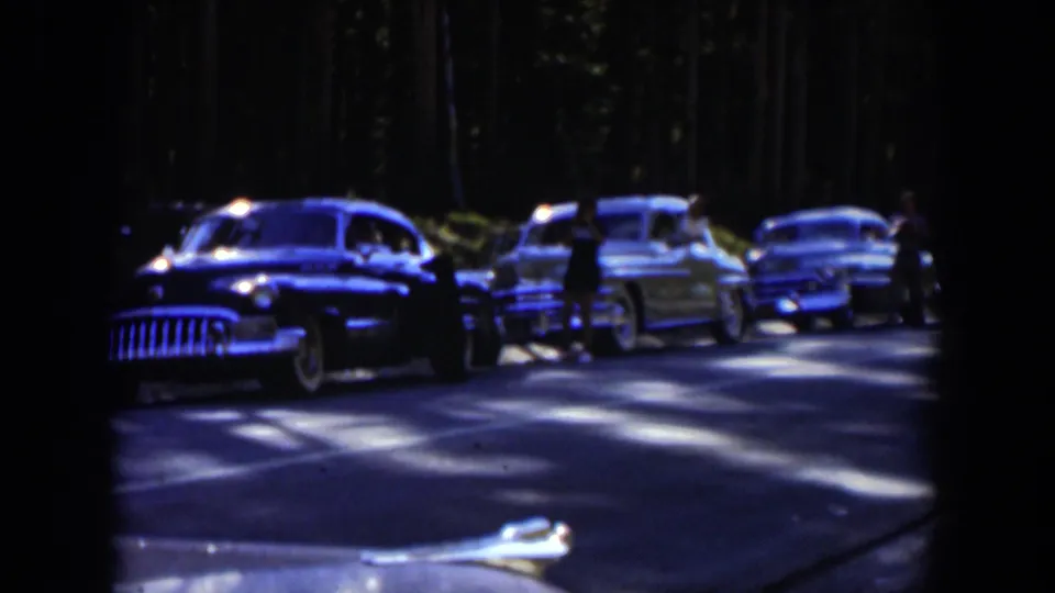 Several mid-century cars sit or move slowly on the shaded Wyoming forest road, with a person possibly near one vehicle.