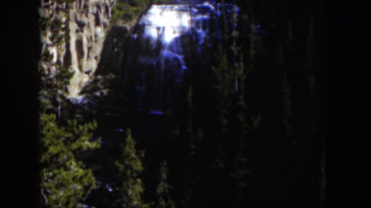 1951 Wyoming 8mm frame of a bright waterfall cascading down rock faces in a shaded evergreen forest.