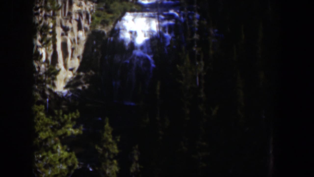 1951 Wyoming 8mm view of a waterfall seen through shadowed trees and pale rock walls.
