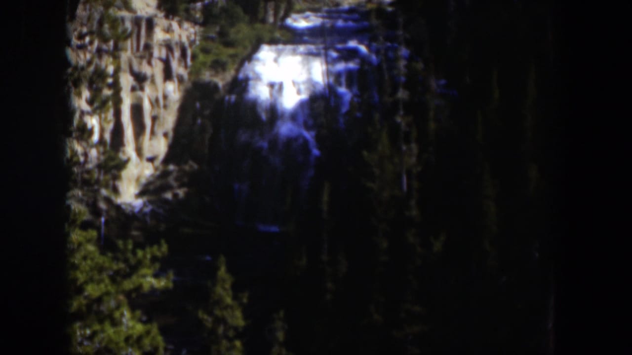 1951 Wyoming 8mm frame showing a bright waterfall over brown rocks, surrounded by dense evergreen forest and deep shadow.