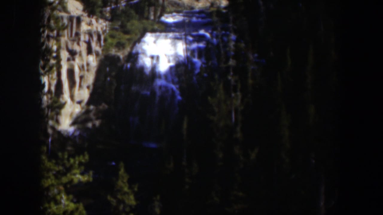 1951 Wyoming 8mm frame with a distant cascade, pale rock face, evergreen trees, and heavy shadow around the falls.