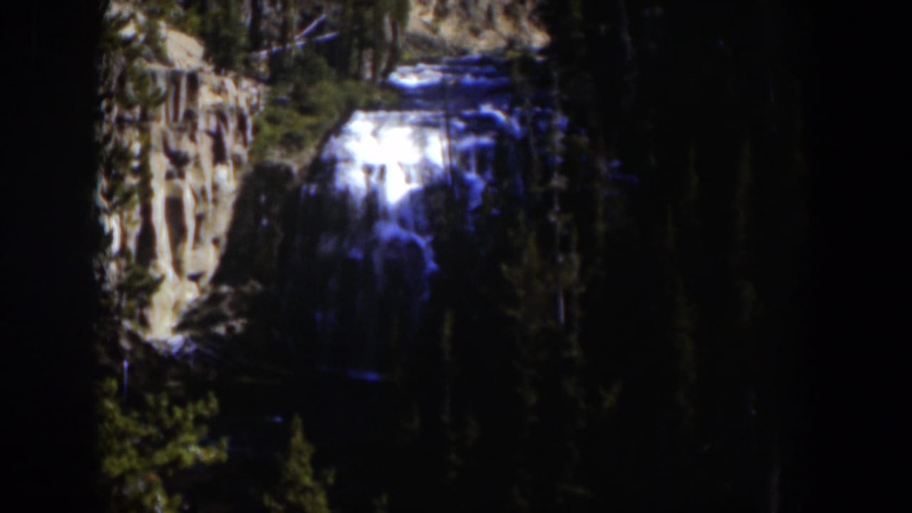 1951 Wyoming home-movie frame showing the waterfall from a distant viewpoint, with brown rocks and dark forest framing the scene.