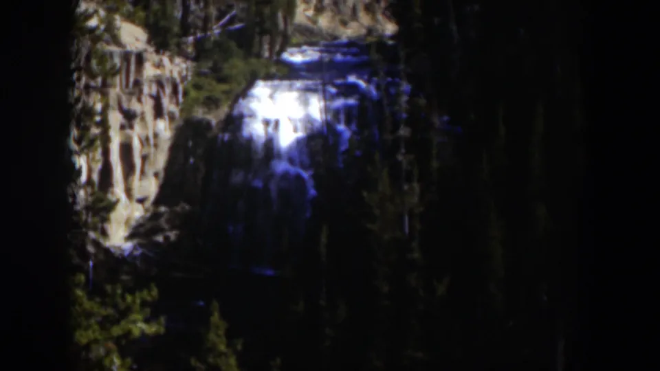 1951 Wyoming home-movie frame showing the waterfall from a distant viewpoint, with brown rocks and dark forest framing the scene.